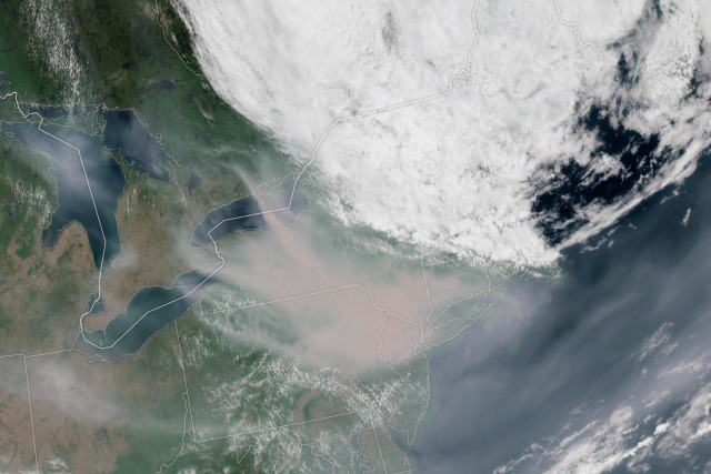 Satellite image of smoke over the northeastern United States. The smoke is a light gray, cottony blanket creating an irregular shape over the center of the image. Behind it, the land surface is light browns and greens. Credit: NASA’s Earth Observatory