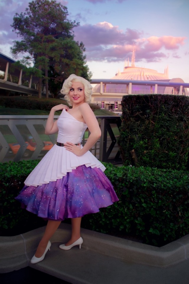 a photo of a woman with blonde hair wearing a dress inspired by space mountain in front of the ride building for space mountain at magic kingdom. the dress is white, cut assymetrically, with stiff pleats around the skirt, and a purple under skirt covered in a galaxy print and rhinestones.