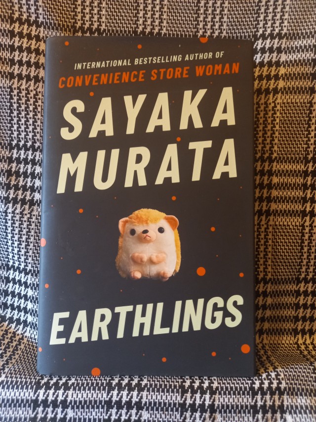 the cover of Earthlings by Sayaka Murata which is black with a cute hedgehog plushie accompanied by red dots The title is in bold white writing across the front of the book. The book rests on a black and white tartan print. 