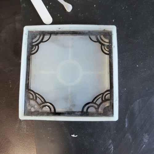 A small, square silicone mold has resin and a layer of paper with metallic swirls of glittery mica powder dusted irregularly around a frame of semi-circles in the corners creating a frame.