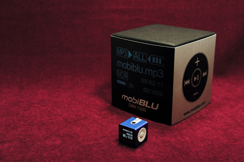 Red background. Image of a black box with white text "MobiBlue MobiBLU DAH-1500i" and an image of the mp3 function buttons on the right side of the box. In front of the box is the MobiBlue MobiBLU DAH-1500i device in the color blue. It is in the shape of a cube. There are white function buttons and the MP3 player appears to be on, with blue digital text against an LSD screen.