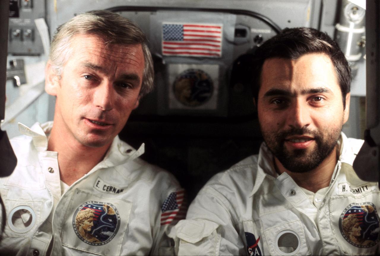 Astronaut Gene Cernan (left) and scientist-astronaut Harrison Schmitt wear white flight suits with Apollo patches on the left chest. Behind them is a gray metal hatch decorated with a small American flag. Credit: NASA