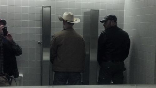 Guy takes a a photo of a cowboy & a biker taking a piss at the urinals