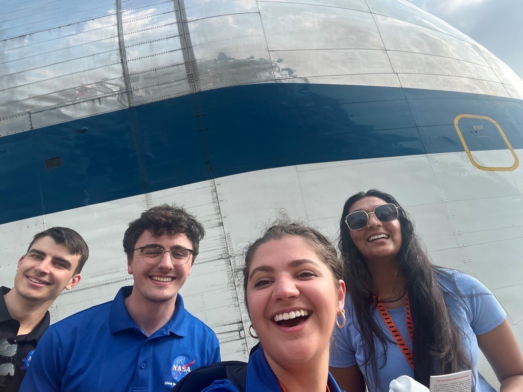 Four people pose in front of NASA’s Super Guppy, a large, specialized aircraft that is used to transport oversize cargo. Each of the four people are wearing a polo shirt with a NASA insignia on the upper left of their shirt. The group is smiling and laughing for the photo. The Super Guppy is shiny and has silver covering the top half of the aircraft, white on the bottom half of the aircraft, and a large blue stripe running along the middle. Credit: NASA