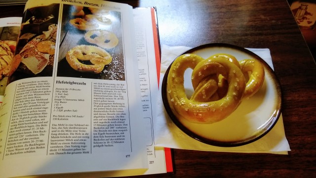 a photo of homemade soft pretzels next to a cookbook open to the recipe page for those pretzels. the recipe is written in german