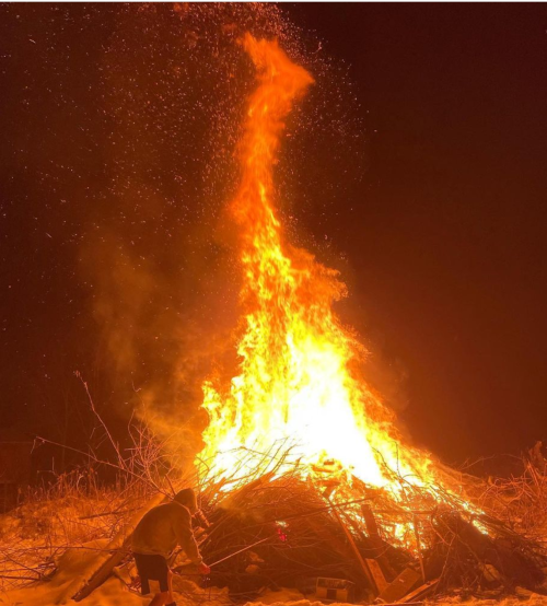 [ID: A roaring bonfire burning at night in a snow-covered field. The pile itself is a bit taller tha