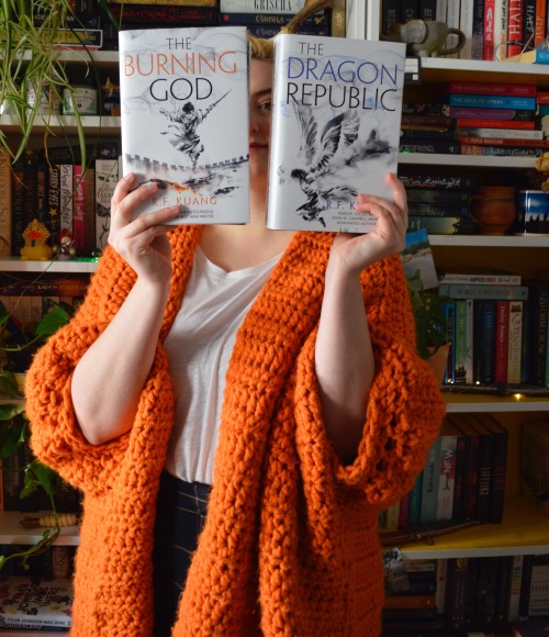 me, standing in front of my bookshelves, holding the burning god and the dragon republic in front of my face with their covers to the camera