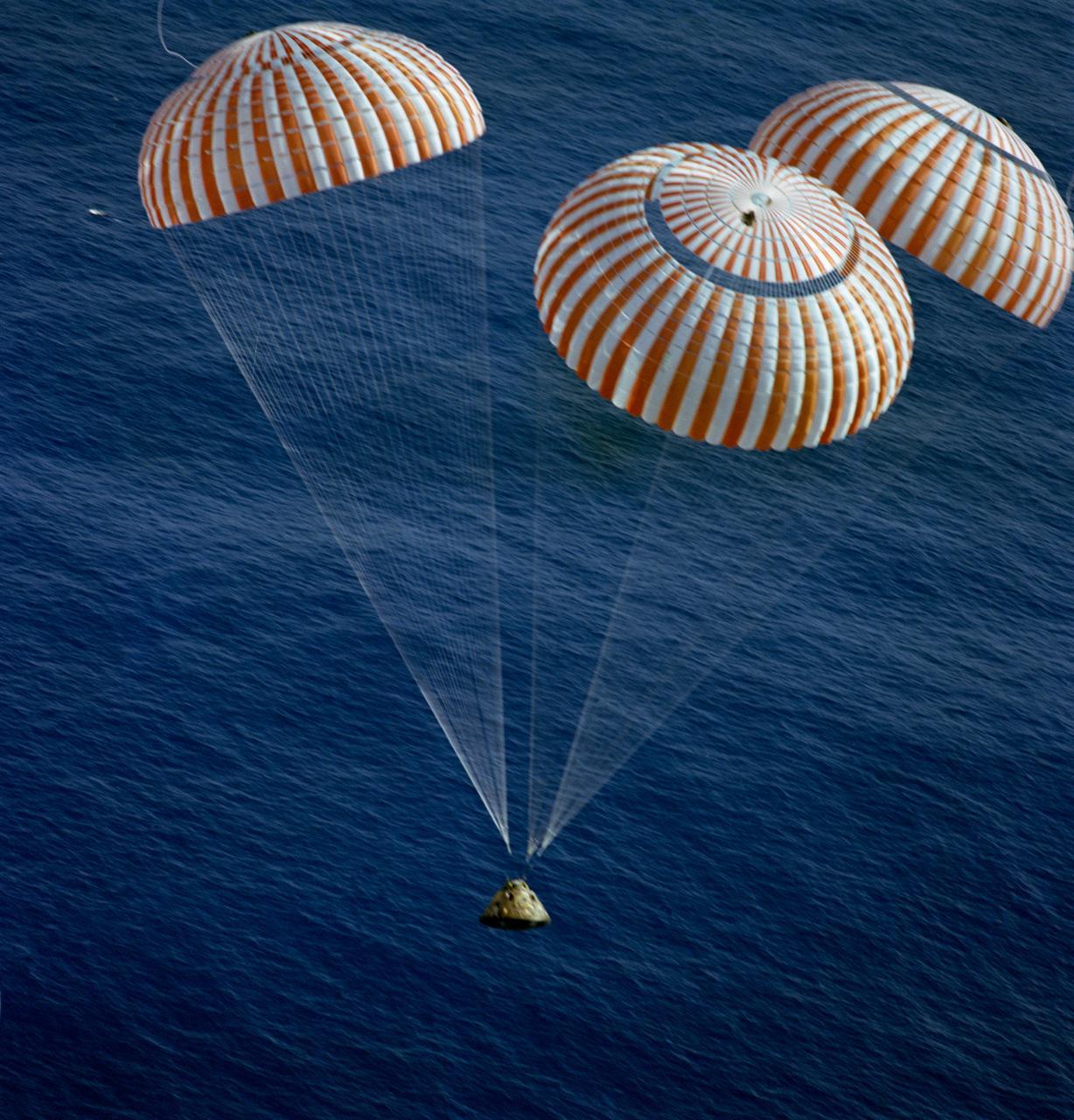 The Apollo 17 Command Module (CM), with astronauts Gene Cernan, Ron Evans and Harrison Schmitt aboard appears as a small conical spaceship.The capsule nears splashdown in the South Pacific Ocean with three enormous red-and-white striped parachutes. This overhead view was taken from a recovery aircraft seconds before the spacecraft hit the blue water. Later, the three crewmen were picked up by a helicopter from the prime recovery ship, USS Ticonderoga. Credit: NASA
