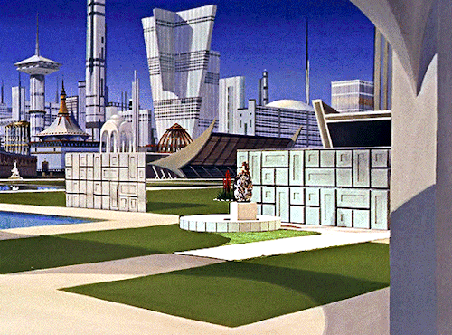 GIF 5: scenic view of a public square and a cityscape (similar but not identical to the first gif). the square itself is several rectangular patches of grass, surrounded by a white sidewalk. on it is, again, a statue and behind it a white wall. on the right is what could be a bridge pier? and the background is a beautiful city skyline with numerous oddly shaped and differntly sized buildings. all the buildings are white. the sky is clear and blue.