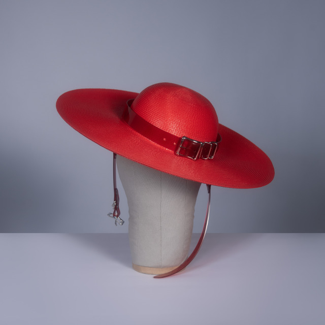 Wide brim hat in red faux straw with translucent red pvc buckle hat band and harness strap details, displayed on a mannequin head against a gray background.