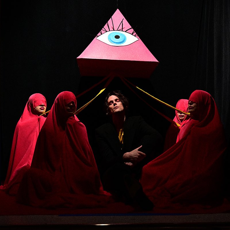 An image of Dallon Weekes sitting cross legged beneath a trianglular prism with an image of an eye on it. He is wearing a black suit and the same star shirt, his head is tilted upwards and his eyes are closed. Surrounding him are four figures wearing red fabric that covers their entire body save for their mouths