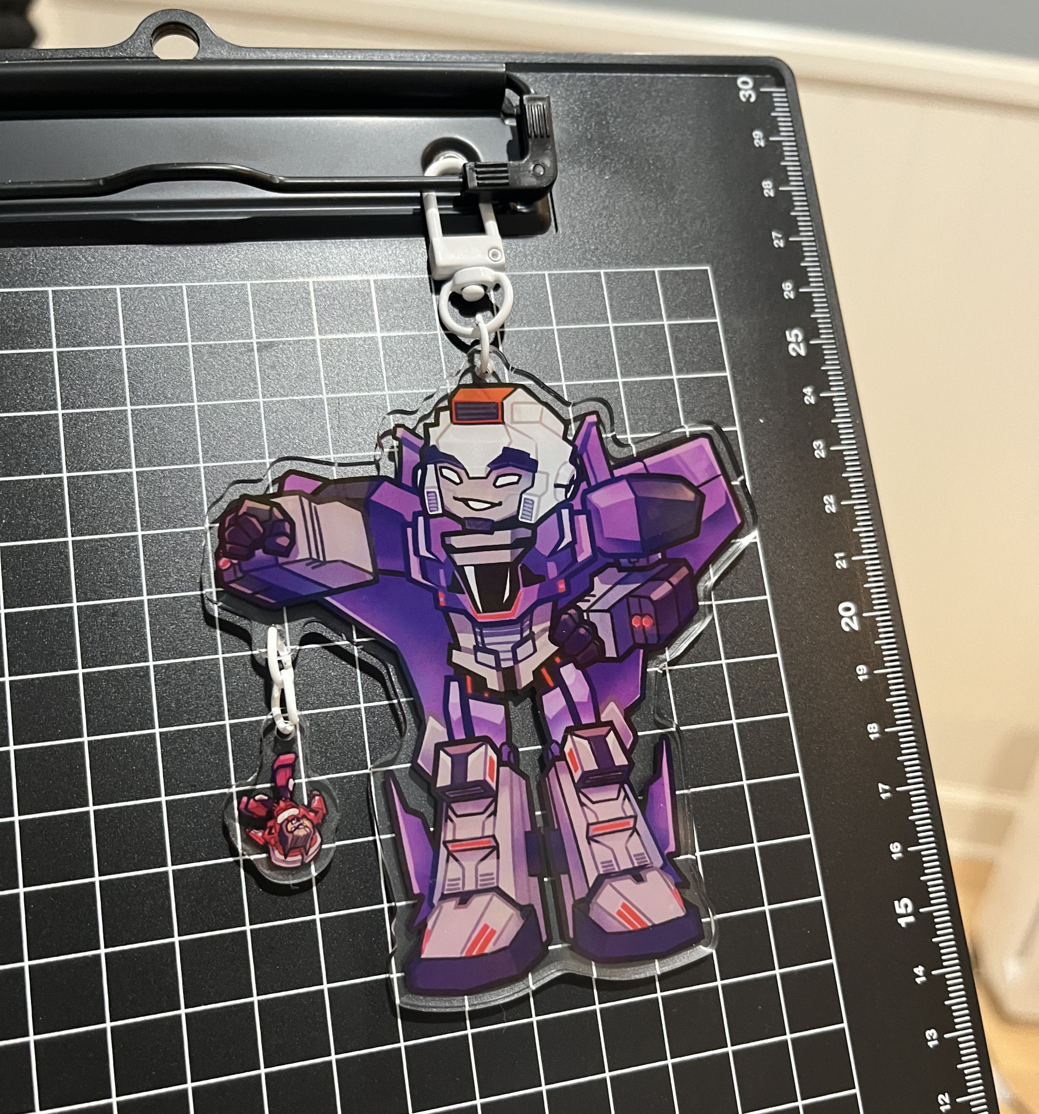A photo of an acrylic charm featuring Astrotrain and Dead End from Transformers: Cyberverse clipped onto a clipboard. Astrotrain is holding Dead End upside down by the foot.
