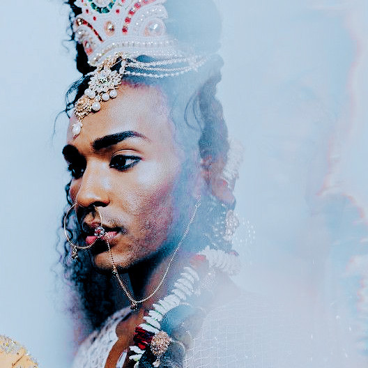 a dark skinned south asian model with braided hair in the backstage of a fashion show. they are wearing kohl and elaborate jewellery including a headdress, hair ornaments and a jewelled naath.