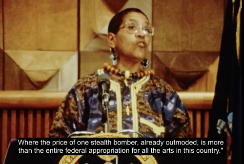 Audre Lorde stands at a podium accepting her award and appointment as State Poet of New York for 1991-1993. She is dressed formally in a colorful tunic. She continues, "'Where the price of one stealth bomber, already outmoded, is more than the entire federal appropriation for all the arts in this country?'"