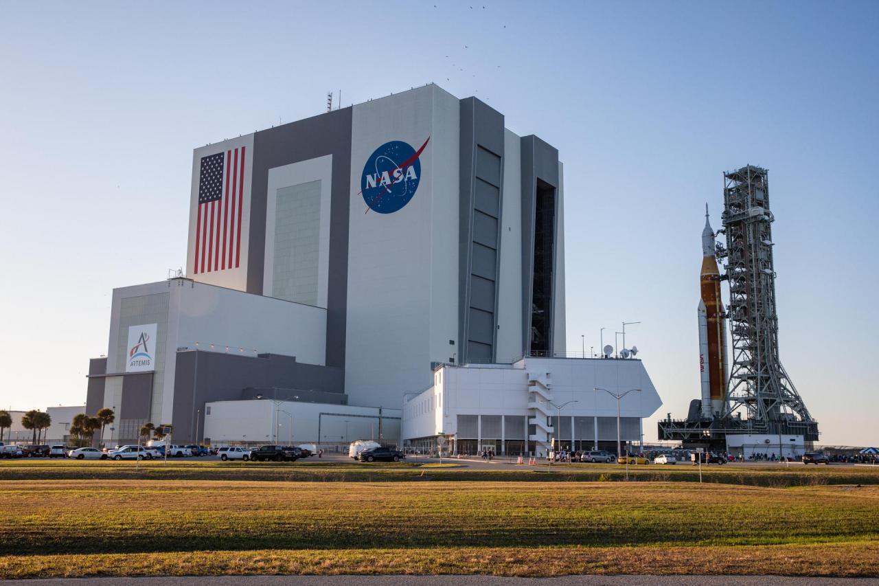 The massive Vehicle Assembly Building (VAB) is a large grey and white cube-shaped building with large doors. It has an American flag on it, along with the NASA meatball logo and the Artemis mission logo. The Space Launch System (SLS) rocket stands to the right of the VAB. The SLS is orange with two white boosters on either side.  Credit: NASA/Kim Shiflett