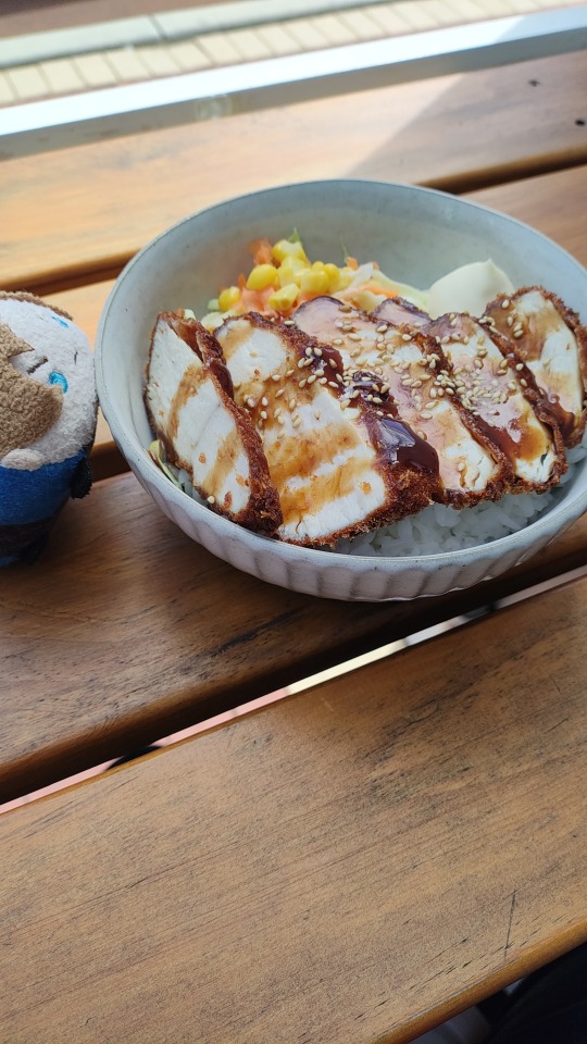Resident Evil 2 Tsum Leon plush toy by a bowl of Katsu chicken with rice, drizzled with Japanese BBQ sauce and a side of lettuce/carrot.