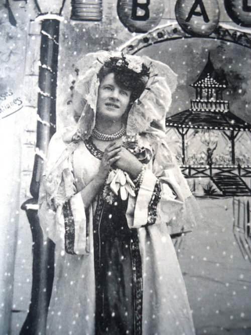 Lovely postcard of woman in her Edwardian coat with hood, who stands at the entrance of a “Ball Masq