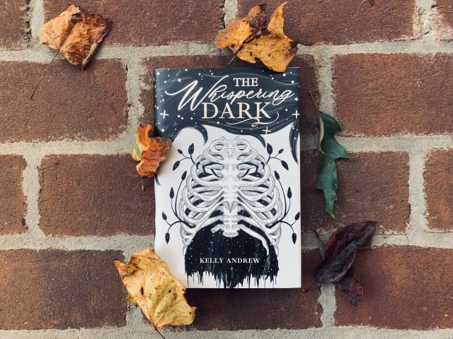 Pictured is the Owlcrate special edition of The Whispering Dark (with black-and-white cover art depicting a skeletal ribcage) against a brick background with autumn leaves surrounding the book. Photo by AHS.