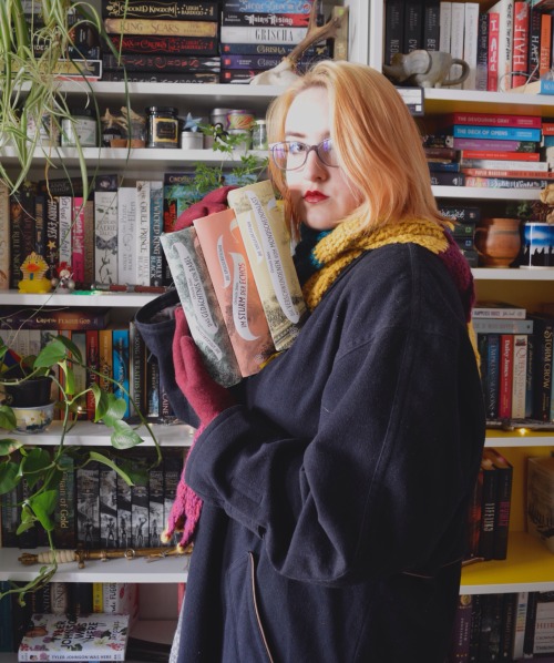 me, standing sideways in front of my bookshelves, holding three books with the spine to the camera in front of me