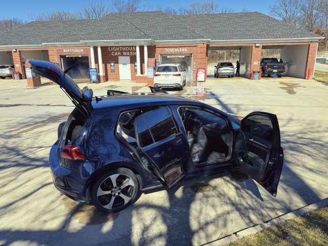 A blue hatchback car with its doors open in the foreground, parked at a car wash facility with "SOFT TOUCH" and "TOUCHLESS" service bays in the background.