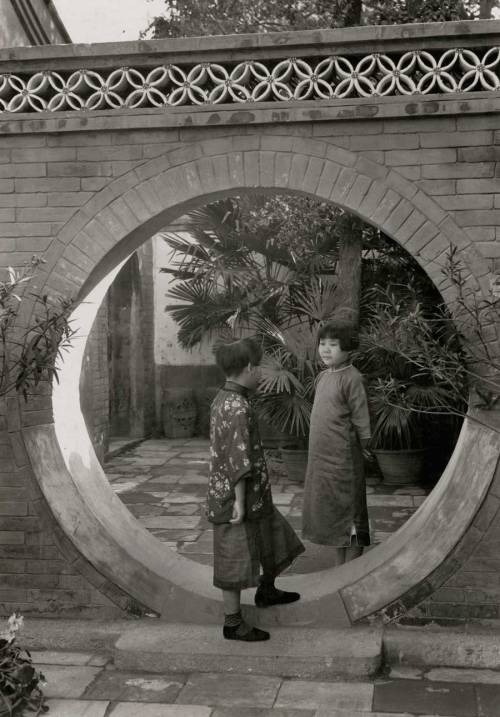 Two young Chinese girls chat with each other in a moon gate in a courtyard of a Chinese home, 1932.P