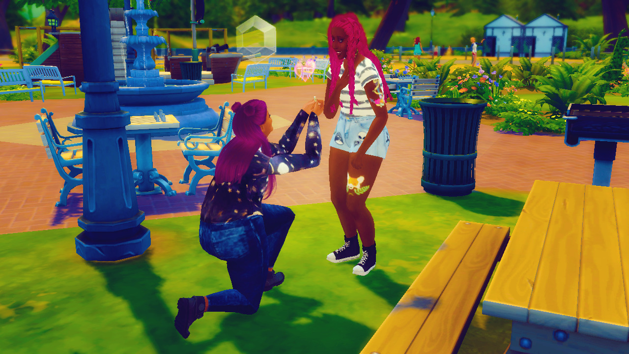 In a park with a fountain, plants, playgrounds, etc. are two women from The Sims 4. On the left  is a brown woman with long purple hair and space buns. She's wearing a moon and sun long sleeve top, jeans, and black boots. She's on one knee with a ring being shown to the other woman and a heart floats over it. The other woman is a black woman with long pink braids, a striped short sleeve shirt, and light shorts with aliens on them. She has a fox tattoo on her left leg and a flower tattoo on her right upper arm. She has one arm over her heart and looks taken by the marriage proposal.