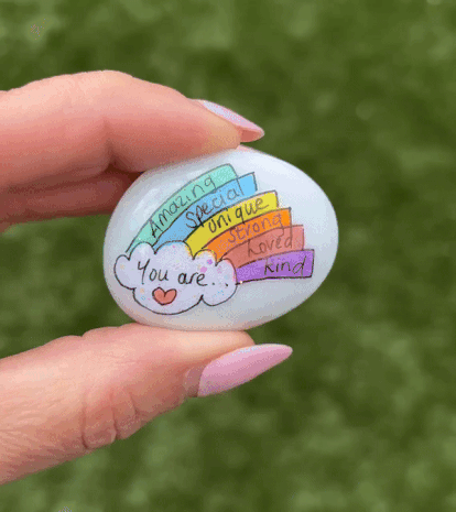 someone holding a white painted rock between their fingers. painted on the rock is a doodle of a rainbow with writing on a cloud and each of the rainbow's stripes. on the cloud is written "you are...", and in order, the words "amazing," "special," "unique, "strong," "loved," and "kind" are written on the stripes.