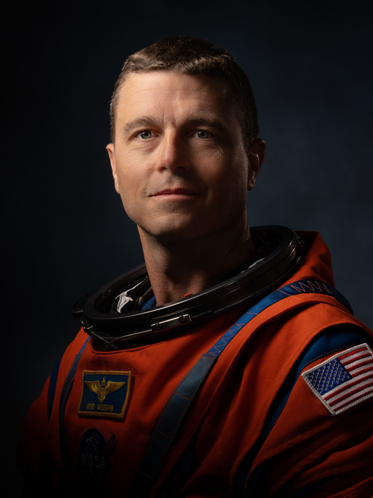 NASA astronaut Reid Wiseman poses for a portrait in his orange Artemis flight suit. The suit has blue trim around the neck and shoulders, with three patches: one with the U.S. flag on his left shoulder, one with his name and a pair of wings on his chest, and one with the NASA “meatball” insignia faintly visible beneath the second. The background is dark, and the photo is lit to focus on Wiseman's face, which is facing the camera with a dignified expression. Credit: NASA