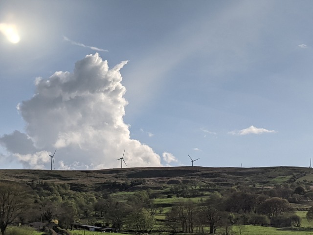 Fields and gentle hills. Wind turbines on the hill, silhouetted against a hazy blue sky. A very very big, very solid white cloud rises up from behind the hill.