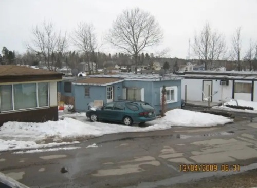 A picture of a trailer park. The trailer to the left is cream-coloured & brown, the one in the center is light blue, & the one to the right is white with black accents. Outside of the blue one is a dark green car, probably from the 1990s. There is melting snow on the ground. The sky is grey. A timestamp in the lower right corner reveals the photo was taken on April 13th 2009 at 6:31am.