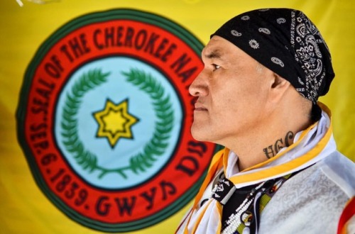 Joshua Squirrel poses for a photo in front of the Cherokee flag, showing off a tattoo of his family 