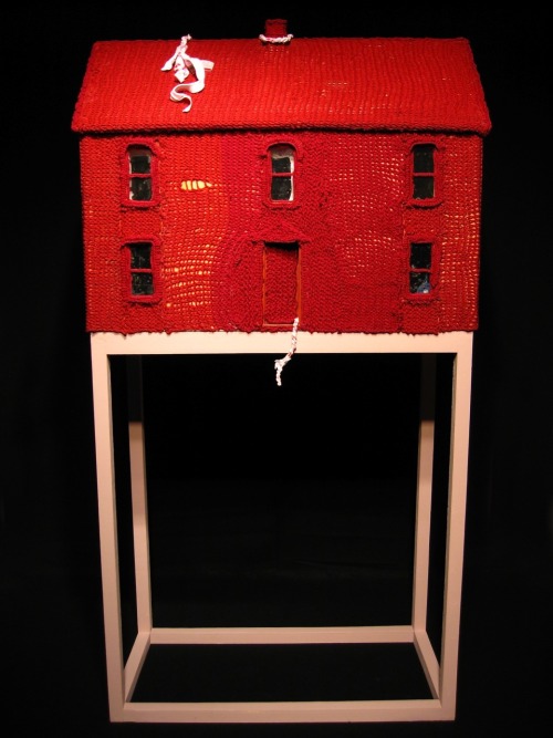 An image of a 3D artpiece called My House by Loren Siems. Against a black backdrop, a constructed model of a house sits on top of a cubic wood frame. The model is symmetrical and the front of the house faces the viewer. There is one door, two windows on either side, and three more identical and evenly spread windows on the second floor. The model is covered in a layer of knitted vivid red yarn, creating the impression that the house is made of knitted red yarn. The windows are dark but there appears to be cluttered objects in the house. A white string is caught in the bottom of the red door. Another string is attached to the chimney at the center of the roof.