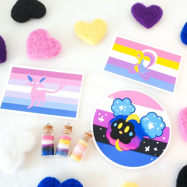 a photo of genderfluid, bigender, and pangender themed items including needle felted hearts, three stickers, and three bottle charms. The stickers feature the pokemon Espeon, Cosmog, and Cresselia.