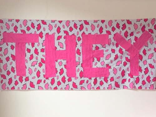 THEY. [rectangular fabric banner that reads they in large pink letters over a pattern with red &