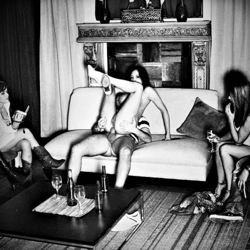Sexplictly erotic photo of a young woman having anal sex on the couch. The two other women -the one at te left reading, the one at the right watching holding a glass of wine- add to the quasi-casual atmosphere. 