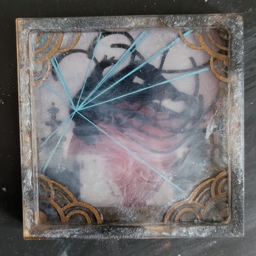 A front view of the craft, out of the silicone mold. The silhouette is obscured somewhat by glittery gray and black mica powder and swirls of white ink. The red circle in the back is indistinctly visible behind the buildings. The shimmery copper frame of semicircles in the corners is most distinct in the foreground.