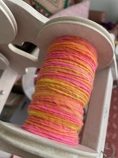 A wooden spinning wheel bobbin, mostly full with handspun neon yarn in orange, yellow, and pink.
