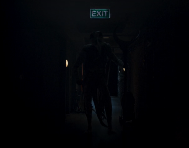 very dark screenshot of a mummified individual shuffling towards the protagonist in a dark hallway. There is a green exit sign above his head