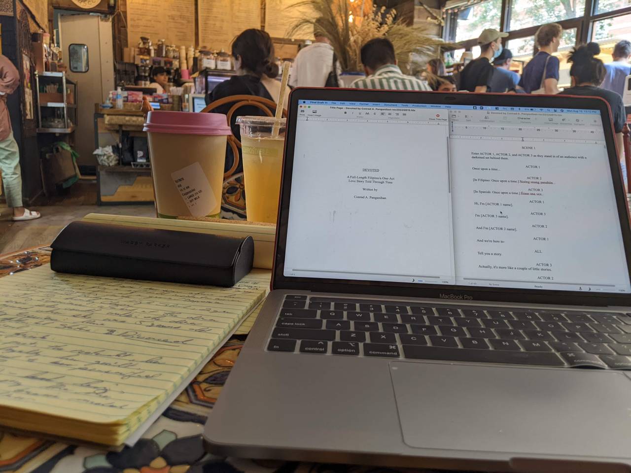 Pictured: a yellow legal pad with a character sketch written in blue ink with an eye glass case on top next to a laptop with the title page for my play, DEVOTED. In the background are two cups: one with coffee and the other is fresh squeezed orange juice and the scene of a busy coffee shop in the Lower East Side, NYC.