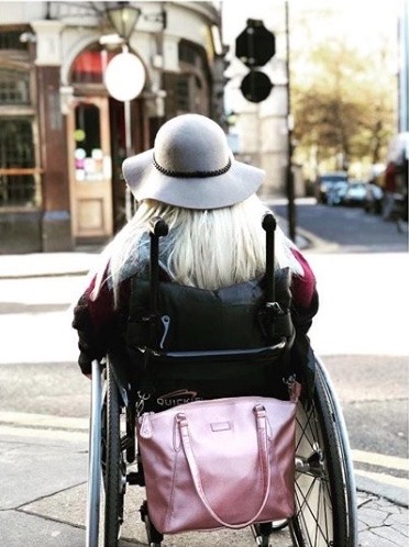 Photograph shows Sam Renke in a wheelchair from the back, with a Rose Gold Samantha Bag stored on the handles using the strap and mobility clips