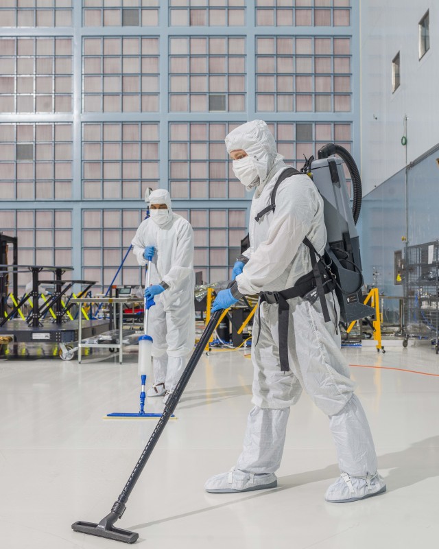 Two people in white “bunny” suits stand on a glossy, white floor. One holds a thin vacuum and the other holds a mop. On the floor behind them are some metallic structures and the wall behind them is covered in pale pink squares. Credit: NASA/Chris Gunn