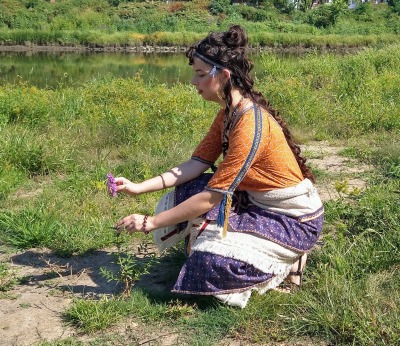 a woman kneels on a riverbank, facing left. The riverbank is covered in bright green grass. The woman is reaching out to touch a tall purple flower growing out of the grass. She wears an orange tunic with blue trim under a beige skirt with two rows each of fringe fringe and blue flounces.