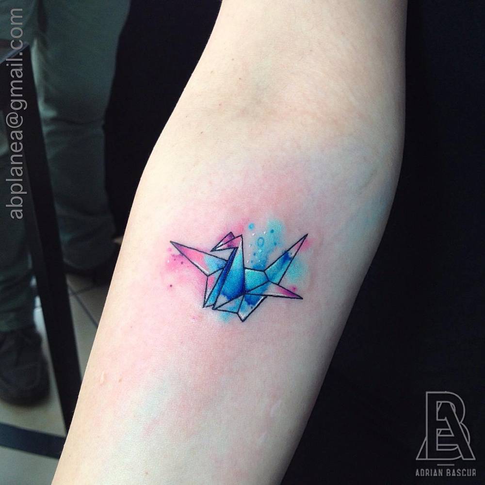 Tatuaje de una grulla de origami con un fondo de acuarela de estilo galáctico situado en el antebrazo. Artista tatuador: Adrian Bascur