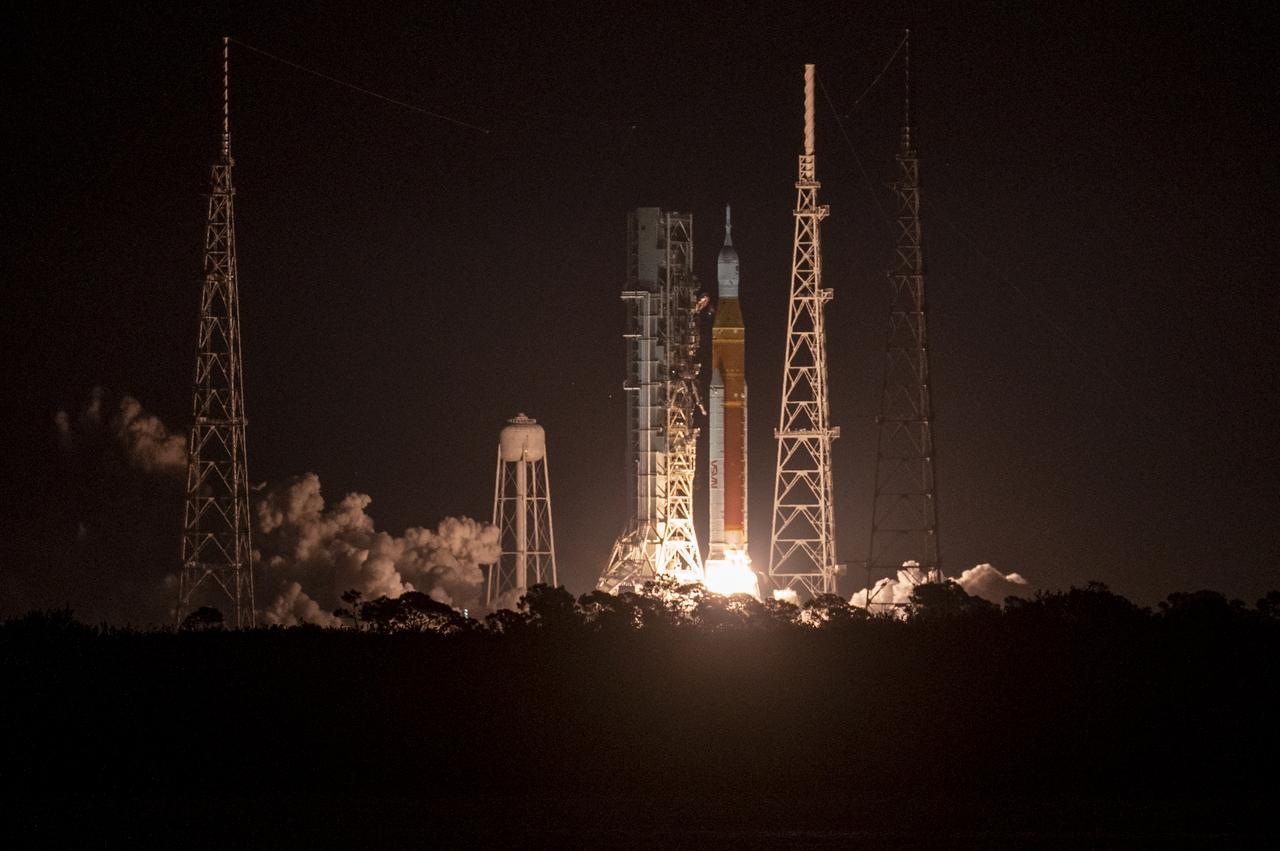 NASA’s Space Launch System rocket carrying the Orion spacecraft launches on the Artemis I flight test, Wednesday, Nov. 16, 2022, from Launch Complex 39B at NASA’s Kennedy Space Center in Florida. The body of the rocket is orange, and it has two white boosters and a white spacecraft sitting on top. As the boosters ignite, they illuminate the launch pad, the water towers, and the lightning towers. The night sky is black in the background. Credits: NASA/Keegan Barber