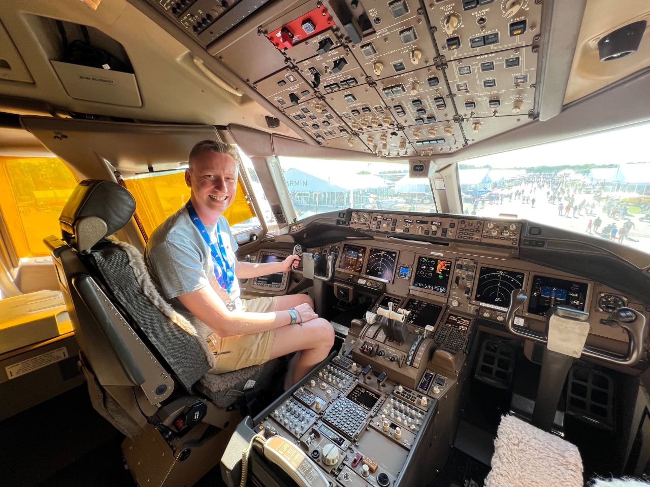 A man wearing a NASA t-shirt sits in a plane cockpit. The cockpit is a metallic grey and covered in hundreds of knobs, throttles, and buttons with two large windows looking forward towards the nose of the plane.