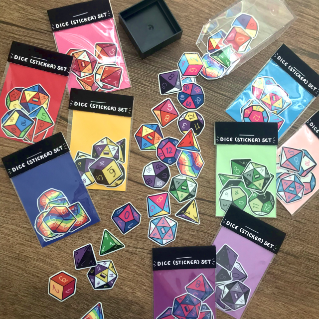 Promotional photo of the previously described dice sticker sets placed around a cascade of individual stickers. These stickers are arranged as though they are tumbling out of an open, plastic Chessex dice container.