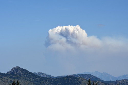 A tall plume of smoke rising above a layer of smoke haze above distant mountain range. 