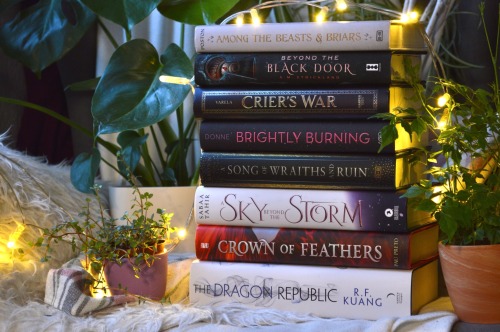 stack of books, surrounded by plants and fairylights, the books are all hardcovers and are from top to bottom: among the beasts & briars, beyond the black door, crier's war, brightly burning, a song of wraiths and ruin, a sky beyond the storm, crown of feathers, the dragon republic