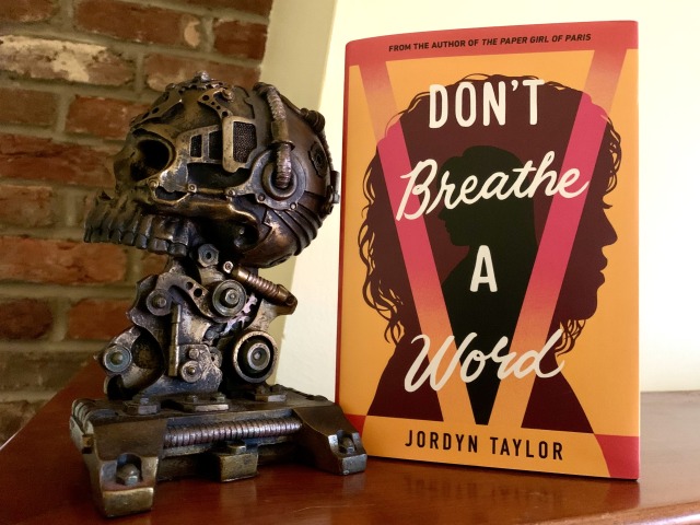 Pictured is DON'T BREATHE A WORD (with cover art depicting one figure in silhouette nested inside another)  set on a mantel, before a brick wall, next to a steampunk skull in profile. Photo by AHS.
