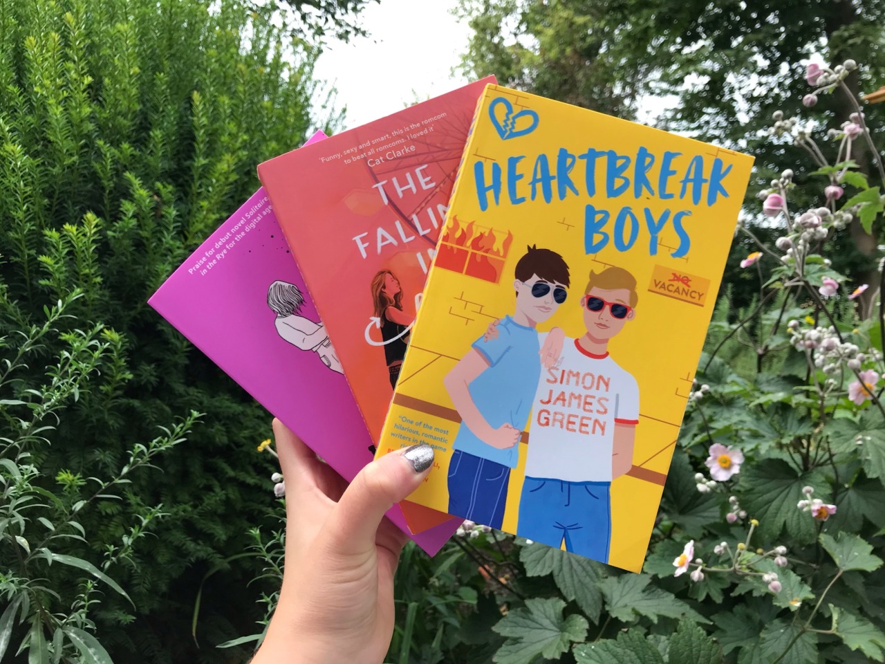 three books shown with their covers towards the camera, heartbreak boys, the falling in love montage behind it and loveless at the end, bushes and flowers in the background
